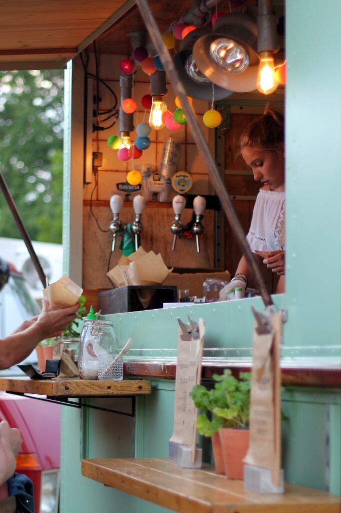Värviliste lampidega kaunistatud toidukaubik, millest valgesse riietunud naine serveerib toitu. Kaubiku ees on näha käed, mis võtavad vastu paberist tuutusse pakitud toitu.